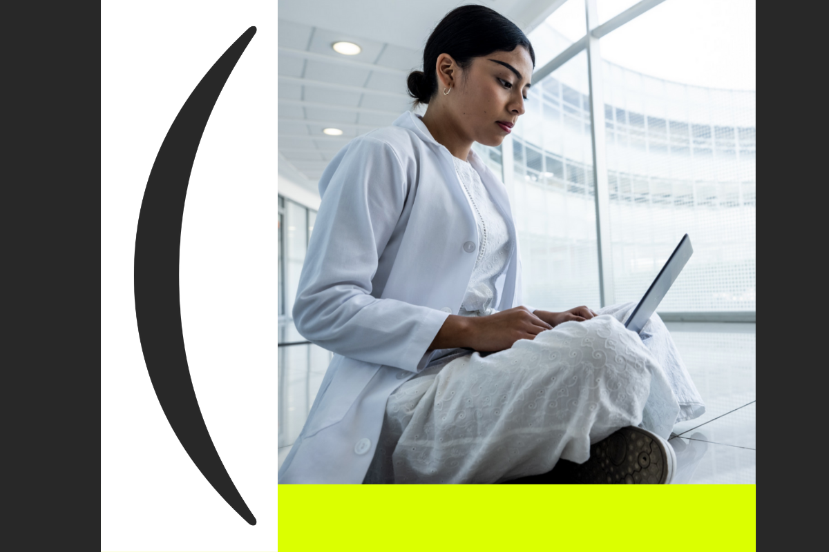 Jóvven estudiante de medicina con bata blancasentada en un pasillo de las instalaciones de la Anáhuac Puebla mirando a una laptop.