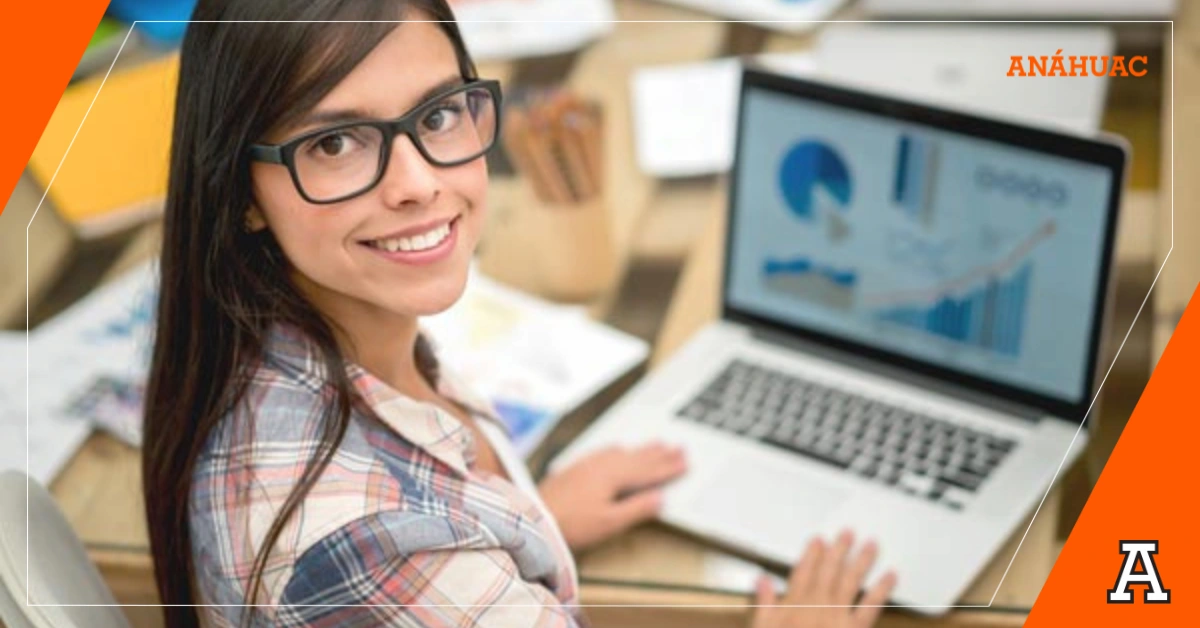 Joven estudiante con lentes volteando a la cámara sonriendo mientras escribe en una computadora con gráficas