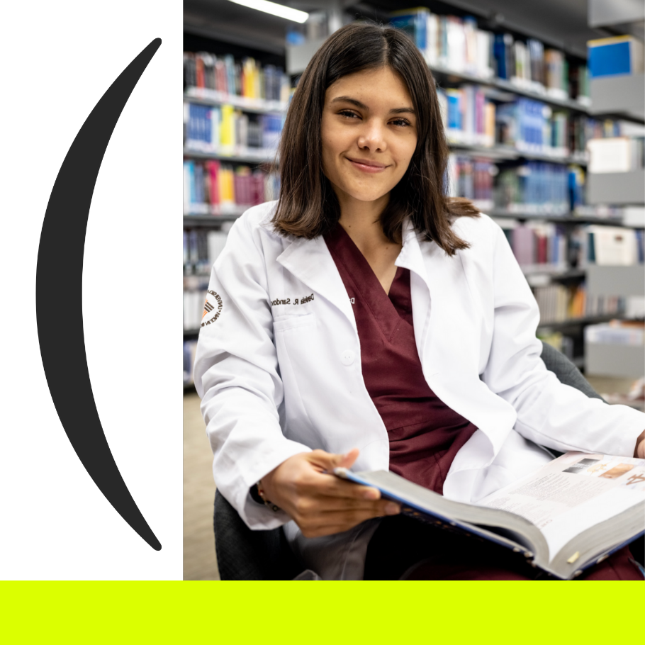 Estudiante del área de salud con bata blanca sentada en la biblioteca de la Anáhuac Puebla leyendo un libro académico. 